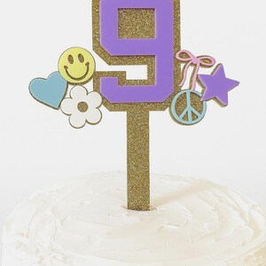 May include: A cake topper with a purple number 9, outlined in gold glitter, adorned with a smiley face, heart, flower, peace sign, star, and bow. The topper is on a cake with white frosting.