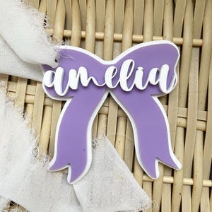 Può includere: Un'etichetta con nome a forma di fiocco viola con il nome "amelia" in corsivo bianco. L'etichetta ha un bordo bianco ed è posta su uno sfondo intrecciato con un accento di tessuto bianco.