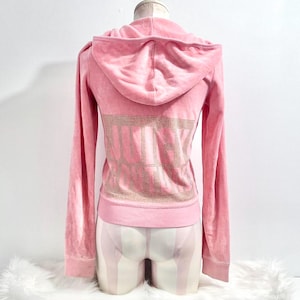 Puede incluir: Una sudadera con capucha y cremallera de terciopelo rosa claro, con capucha y mangas largas. La parte trasera de la sudadera tiene la palabra "JUICY" escrita con pedrería dorada. La sudadera se muestra en un maniquí.