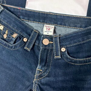 Puede incluir: Primer plano de unos vaqueros de mezclilla azul oscuro con herrajes dorados. Los vaqueros tienen un dise&ntilde;o cl&aacute;sico de cinco bolsillos, cierre de bot&oacute;n y una etiqueta que dice "MADE IN USA 26".