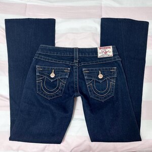 Puede incluir: Vaqueros de mezclilla azul oscuro con pernera acampanada. Los vaqueros presentan un dise&ntilde;o cl&aacute;sico de cinco bolsillos con costuras decorativas y herrajes dorados. Un parche de la marca True Religion es visible en la cinturilla trasera.