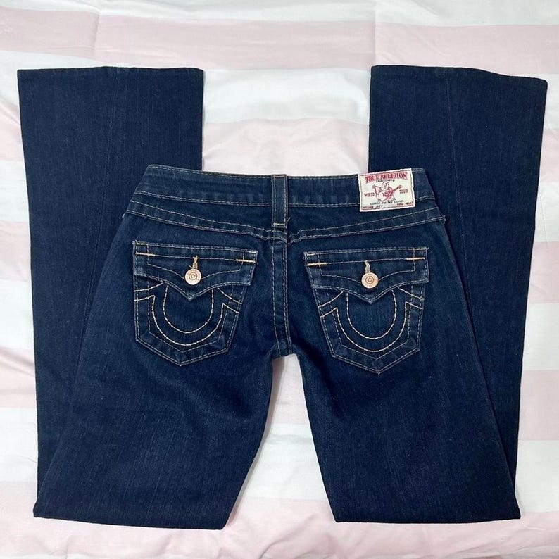 Puede incluir: Vaqueros de mezclilla azul oscuro con pernera acampanada. Los vaqueros presentan un dise&ntilde;o cl&aacute;sico de cinco bolsillos con costuras decorativas y herrajes dorados. Un parche de la marca True Religion es visible en la cinturilla trasera.