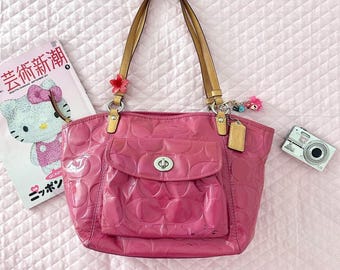 May include: A bright pink patent leather tote bag featuring a debossed signature pattern, a front flap pocket with a silver-tone turn-lock closure, and tan leather shoulder straps. The bag includes a small pink flower charm and a metal tag.