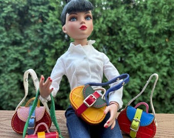 May include: A doll wearing a white blouse and blue jeans, holding several miniature, colorful handbags. The handbags are red, yellow, blue, and pink, with contrasting straps and buckles. The doll has black hair and red lipstick.