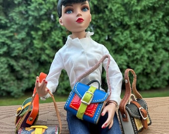 May include: A doll wearing a white blouse and blue jeans, with three miniature handbags in different colors and patterns. The handbags have leather straps and buckles. The doll has black hair and red lipstick.