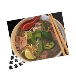 以下が含まれることがあります： ベトナムの麺スープ、フォーのボウル。箸と赤いスプーンが添えられています。スープには麺、肉、もやし、ハーブ、ライムが入っています。画像はパズルです。