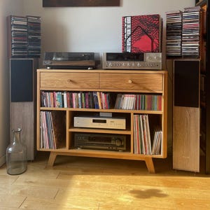 Może przedstawiać: Drewniana szafka rozrywkowa z gramofonem, odbiornikiem stereo i głośnikami. Szafka mieści płyty CD i winylowe. Obraz wisi nad szafką. Przezroczysta szklana butelka stoi na podłodze.