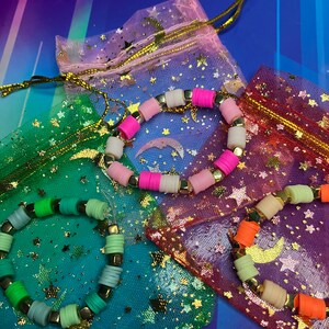 May include: Three colourful beaded bracelets with gold accents, each in a separate star-patterned mesh bag. The bracelets are pink and white, green and blue, and orange and beige.