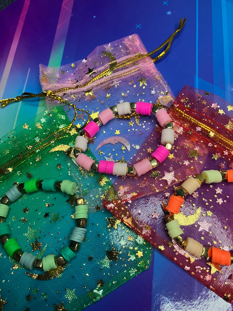 May include: Three colourful beaded bracelets with gold accents, each in a separate star-patterned mesh bag. The bracelets are pink and white, green and blue, and orange and beige.
