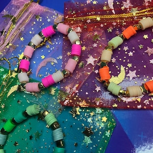 Puede incluir: Tres pulseras de cuentas coloridas con detalles dorados, mostradas en bolsas de tela con estampado de estrellas.