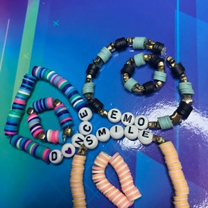 May include: A set of colourful beaded bracelets with the words "DANCE", "SMILE", and "EMO" spelled out in black letters. The bracelets are made of various colours, including pink, blue, yellow, and white.