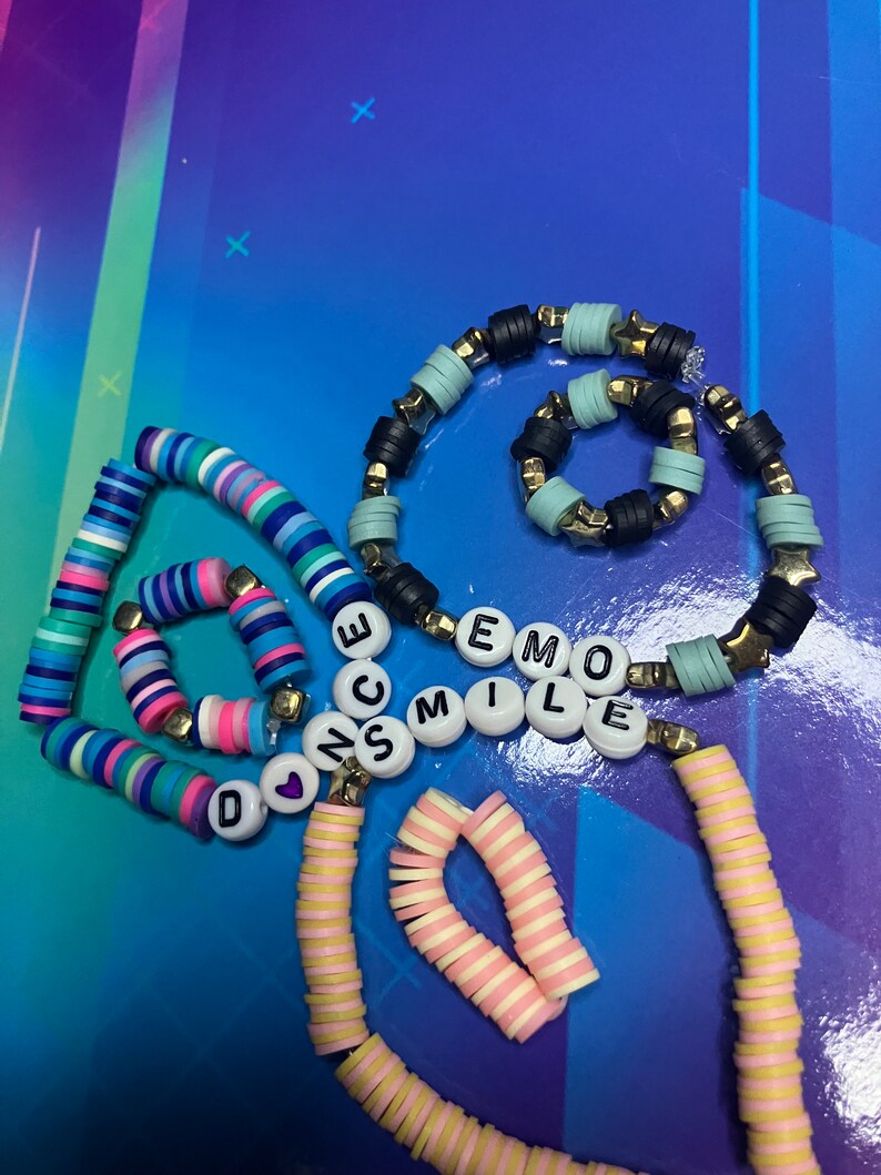 May include: A set of colourful beaded bracelets with the words "DANCE", "SMILE", and "EMO" spelled out in black letters. The bracelets are made of various colours, including pink, blue, yellow, and white.