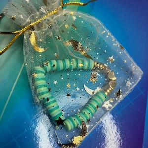 May include: A green and white striped beaded bracelet with gold accents, displayed in a white mesh bag with gold stars and crescent moons.