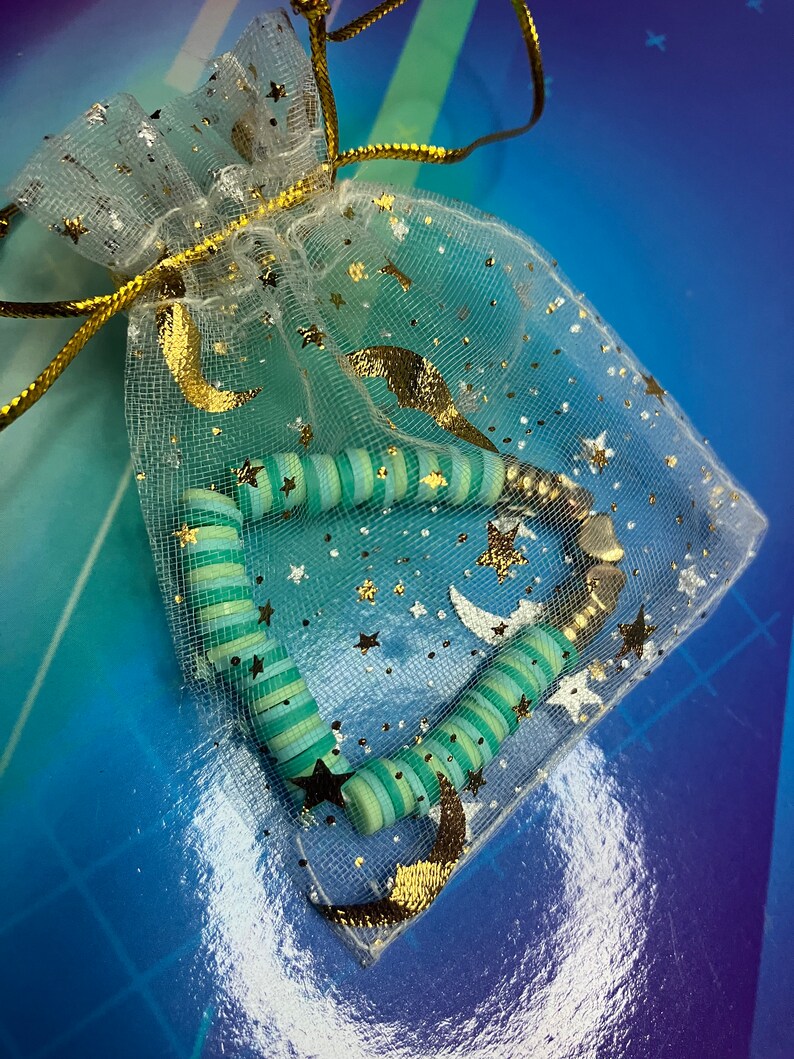May include: A green and white striped beaded bracelet with gold accents, displayed in a white mesh bag with gold stars and crescent moons.