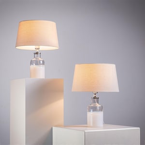 Op de afbeelding: Twee tafellampen met beige stoffen lampenkappen en heldere glazen voet. De voet is wit aan de onderkant en heeft een zilveren metalen bovenkant.