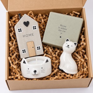 May include: A gift box containing a grey wooden house ornament with a black heart and the word HOME, a sage green square box with the text Home is where the cat is, a white ceramic cat-shaped tea light holder, and a white ceramic cat figurine.