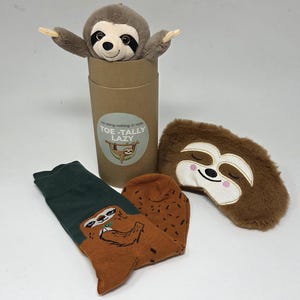 May include: A brown plush sloth toy emerging from a cylindrical cardboard tube with a label reading Toe-Tally Lazy, a pair of dark green and brown socks featuring a sloth graphic, and a brown faux fur sleep mask with an embroidered sloth face.
