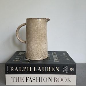 Puede incluir: Una jarra alta de cerámica beige moteada con un asa curva y un pico vertedor. La jarra está colocada sobre dos libros: "Ralph Lauren in His Own Fashion" y "The Fashion Book".