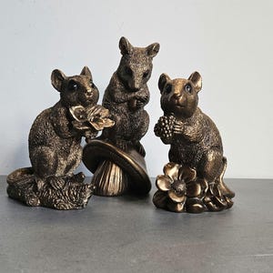 May include: Three bronze figurines of mice, each with a different pose and holding a different object. One mouse is holding a flower, another is holding a mushroom, and the third is holding a cluster of berries.