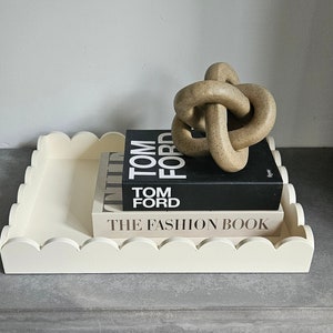 Puede incluir: Una bandeja blanca con bordes festoneados con dos libros apilados encima. El libro de arriba es negro con texto blanco que dice "TOM FORD". El libro de abajo es blanco con texto negro que dice "THE FASHION BOOK". Una escultura anudada marrón se encuentra encima de los libros.