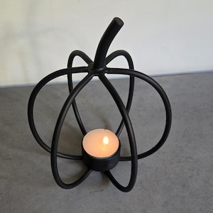 May include: A matte black metal candle holder shaped like a pumpkin with curved wire ribs forming the structure. A small circular base at the centre holds a single lit tea light candle with a warm glowing flame, creating a simple silhouette.
