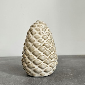 Op de afbeelding: Een witte keramische dennenappel ornament met een getextureerd oppervlak. De dennenappel is een decoratief object voor thuis.
