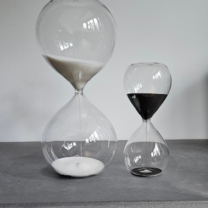 May include: Two glass hourglasses, one large and one small, with white sand in the top bulb of the larger hourglass and black sand in the top bulb of the smaller hourglass.