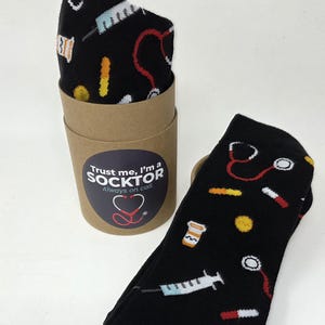 May include: A pair of black socks featuring a medical-themed pattern of stethoscopes, syringes, and pills. One sock is folded inside a cylindrical cardboard sleeve with a label that reads: Trust me, I'm a SOCKTOR Always on call.