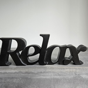 Può includere: Lettere in legno nere che compongono la parola "Relax" su una superficie grigia.