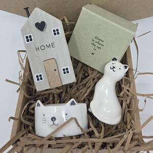 Op de afbeelding: Een geschenkdoos met een lichtgrijs houten huisornament met een zwart hart en het woord HOME, een vierkante lichtgroene doos met de tekst Home is where the cat is, een wit keramieken kattenbeeldje en een wit keramieken kom met kattengezicht.
