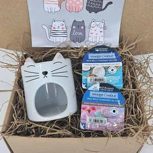 May include: A white ceramic cat-shaped wax melt warmer with a black nose and whiskers. The warmer is in a brown cardboard box with shredded paper. Two Yankee Candle wax melts are also in the box. One is a coconut splash scent and the other is a berry mochi scent.