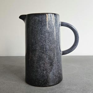Op de afbeelding: Een donkerblauwe keramische kan met een gespikkelde afwerking, handvat en tuit. De cilindrische kan is geschikt voor het serveren van dranken. De hoogte is ongeveer 20 cm.