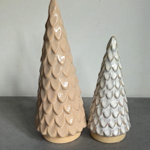 Op de afbeelding: Twee conische keramische sculpturen in de vorm van bomen met een textuur van overlappende schubben. Het hogere beeldhouwwerk is effen lichtbeige, terwijl het kortere beeldhouwwerk wit is met subtiele beige accenten op de randen.