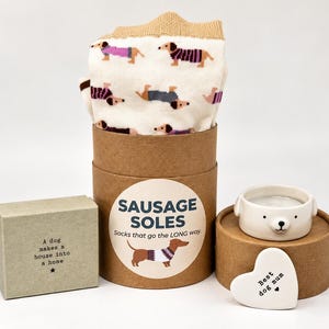 May include: A pair of cream-coloured socks with a dachshund pattern inside a brown cardboard tube labelled Sausage Soles. Beside it sits a small rectangular box with the text A dog makes a house into a home and a white ceramic bear bowl with a heart tag.