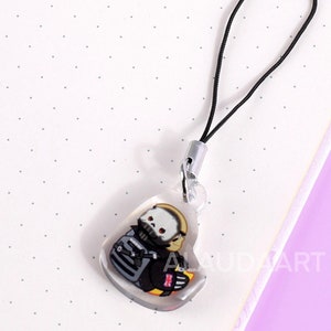May include: A clear heart-shaped charm with a black and white illustration of a character wearing a helmet and backpack. The charm is attached to a black string.