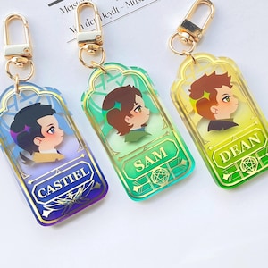 May include: Three acrylic keychains with gold-colored hardware. Each keychain features a cartoon character portrait and a name: Castiel, Sam, and Dean. The keychains have a decorative border and are in shades of blue, green, and yellow.