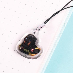 May include: A clear acrylic charm with a black cord. The charm features a colorful illustration of a character in a military-style outfit. The charm is hanging over a white dotted notebook and a light blue surface.
