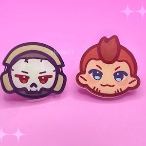 May include: Two circular acrylic pins featuring stylised character illustrations. The left pin shows a white skull with red eyes wearing tan headphones. The right pin depicts a character with reddish-brown hair, blue eyes, and a small mouth.