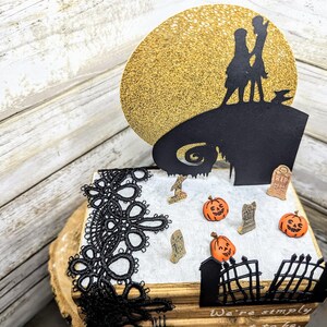 May include: A black and white silhouette of Jack Skellington and Sally from the Nightmare Before Christmas movie on a gold glitter background. The silhouette is on top of a book with black lace trim. The book has a white surface with a black gate, tombstones, and pumpkins.