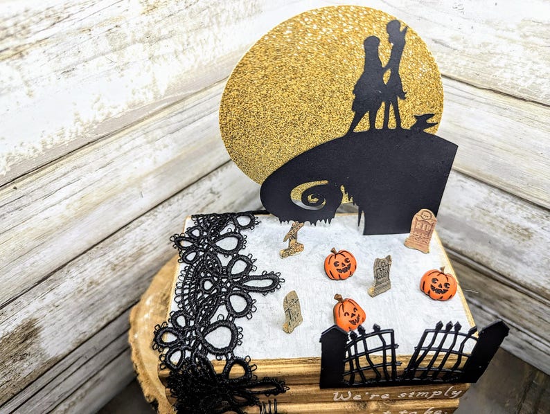 May include: A black and white silhouette of Jack Skellington and Sally from the Nightmare Before Christmas movie on a gold glitter background. The silhouette is on top of a book with black lace trim. The book has a white surface with a black gate, tombstones, and pumpkins.
