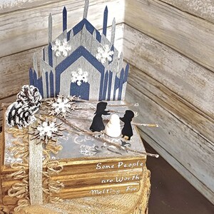 May include: A stack of books with a blue and gray paper castle on top. The books have the text "Some People are Worth Melting For" on the cover. The castle is decorated with white snowflakes.