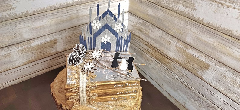 May include: A stack of books with a blue and gray paper castle on top. The books have the text "Some People are Worth Melting For" on the cover. The castle is decorated with white snowflakes.