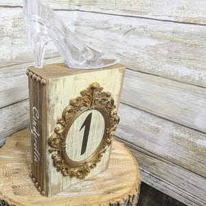 May include: A clear plastic high-heeled shoe sits atop a wooden block table number. The block is painted cream with a gold-colored ornate frame around the number 1. The word "Cinderella" is written on the side.