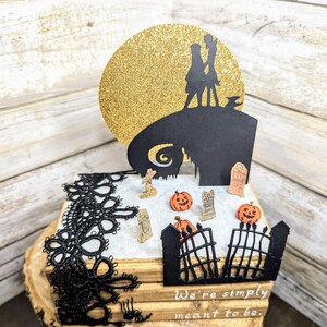 May include: A black and white silhouette of Jack Skellington and Sally from the Nightmare Before Christmas movie, standing on a hill with a gold glitter moon behind them. The scene is on a stack of books with black lace trim and a small gate with pumpkins and tombstones.