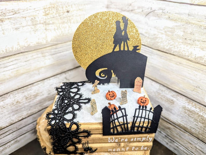 May include: A black and white silhouette of Jack Skellington and Sally from the Nightmare Before Christmas movie, standing on a hill with a gold glitter moon behind them. The scene is on a stack of books with black lace trim and a small gate with pumpkins and tombstones.