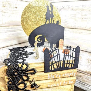 May include: A black and white papercraft scene of Jack Skellington and Sally from the Nightmare Before Christmas movie. The scene is set on a stack of wooden books with black lace trim. The scene includes a golden glitter moon, a graveyard, and a gate. The text "We're simply meant to be" is written on the books.