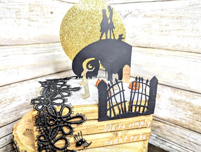 May include: A black and white papercraft scene of Jack Skellington and Sally from the Nightmare Before Christmas movie. The scene is set on a stack of wooden books with black lace trim. The scene includes a golden glitter moon, a graveyard, and a gate. The text "We're simply meant to be" is written on the books.