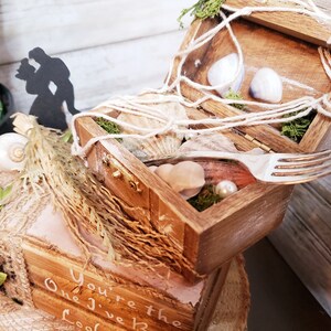 K&ouml;nnte beinhalten: Eine Holzkiste mit einem Seilgriff, ge&ouml;ffnet, um Muscheln, Moos und eine Gabel zu enth&uuml;llen. Die Kiste steht auf einer kleineren Holzkiste mit dem Text "You're the One I Love" darauf.