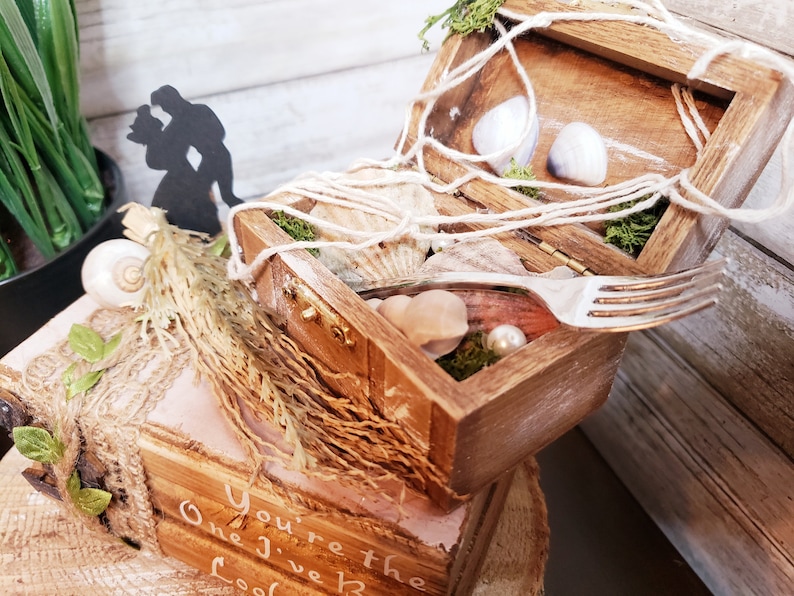 K&ouml;nnte beinhalten: Eine Holzkiste mit einem Seilgriff, ge&ouml;ffnet, um Muscheln, Moos und eine Gabel zu enth&uuml;llen. Die Kiste steht auf einer kleineren Holzkiste mit dem Text "You're the One I Love" darauf.