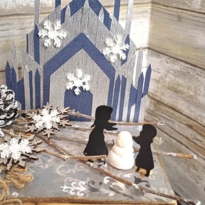May include: A miniature winter scene with a blue and gray paper castle, two black paper figures, a white paper snowman, and brown pine cones on a wooden surface.